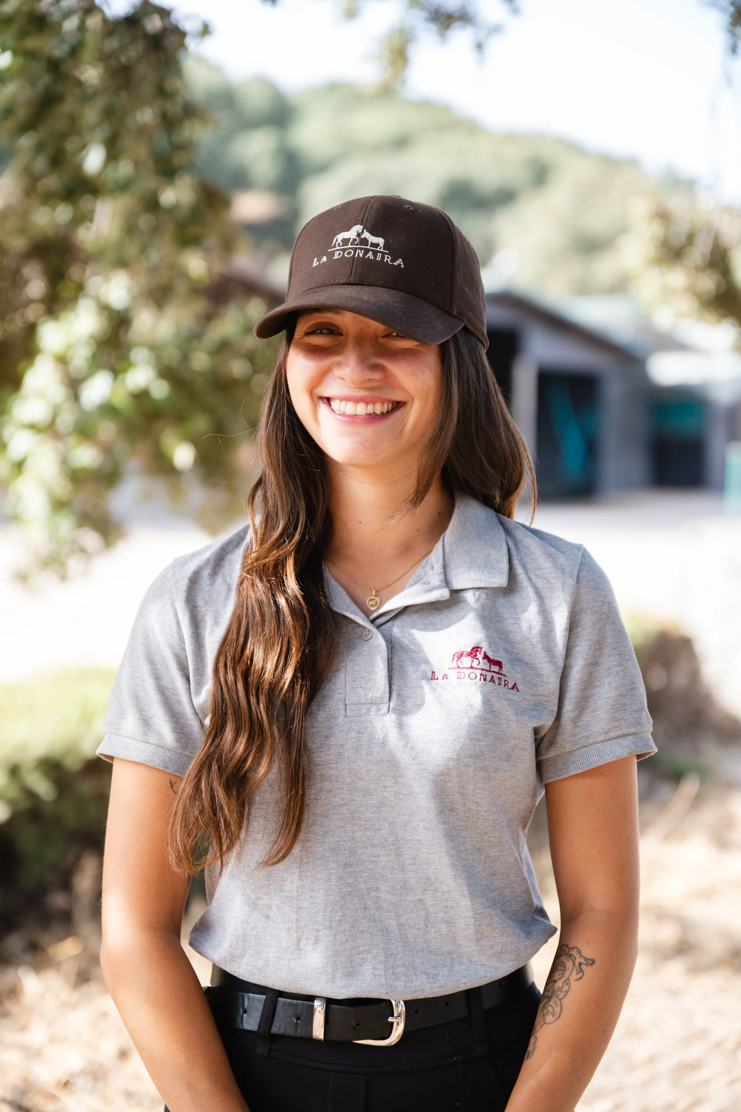 A young woman with blonde hair wearing a black baseball cap and a gray polo shirt with a logo, outdoors in a natural setting with blurred trees and greenery in the background.