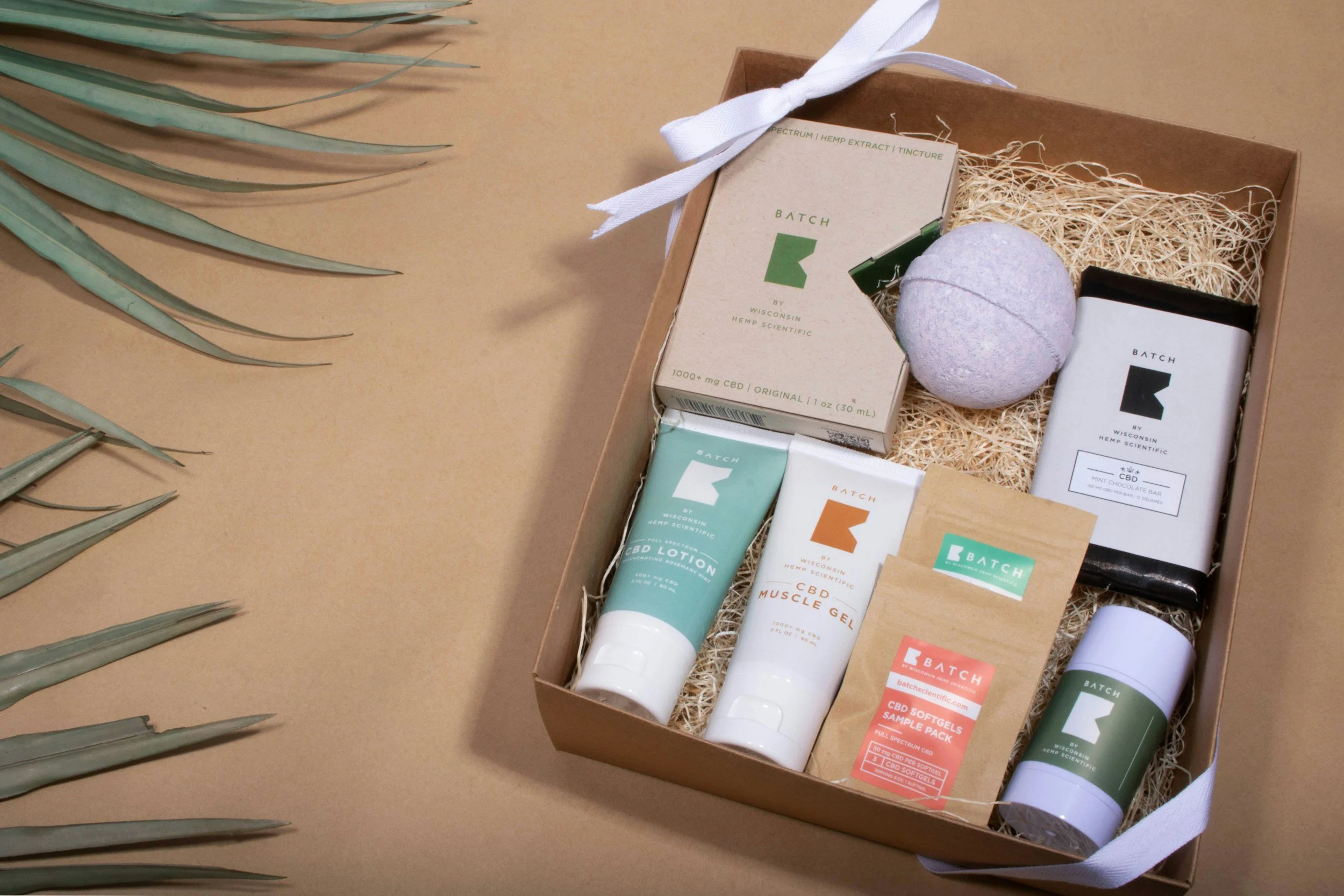 A gift box containing CBD products including CBD lotion, muscle gel, softgels, tincture, and a bath bomb, all arranged on shredded paper with a white ribbon tied around the box, on a beige background with greenery on the side.