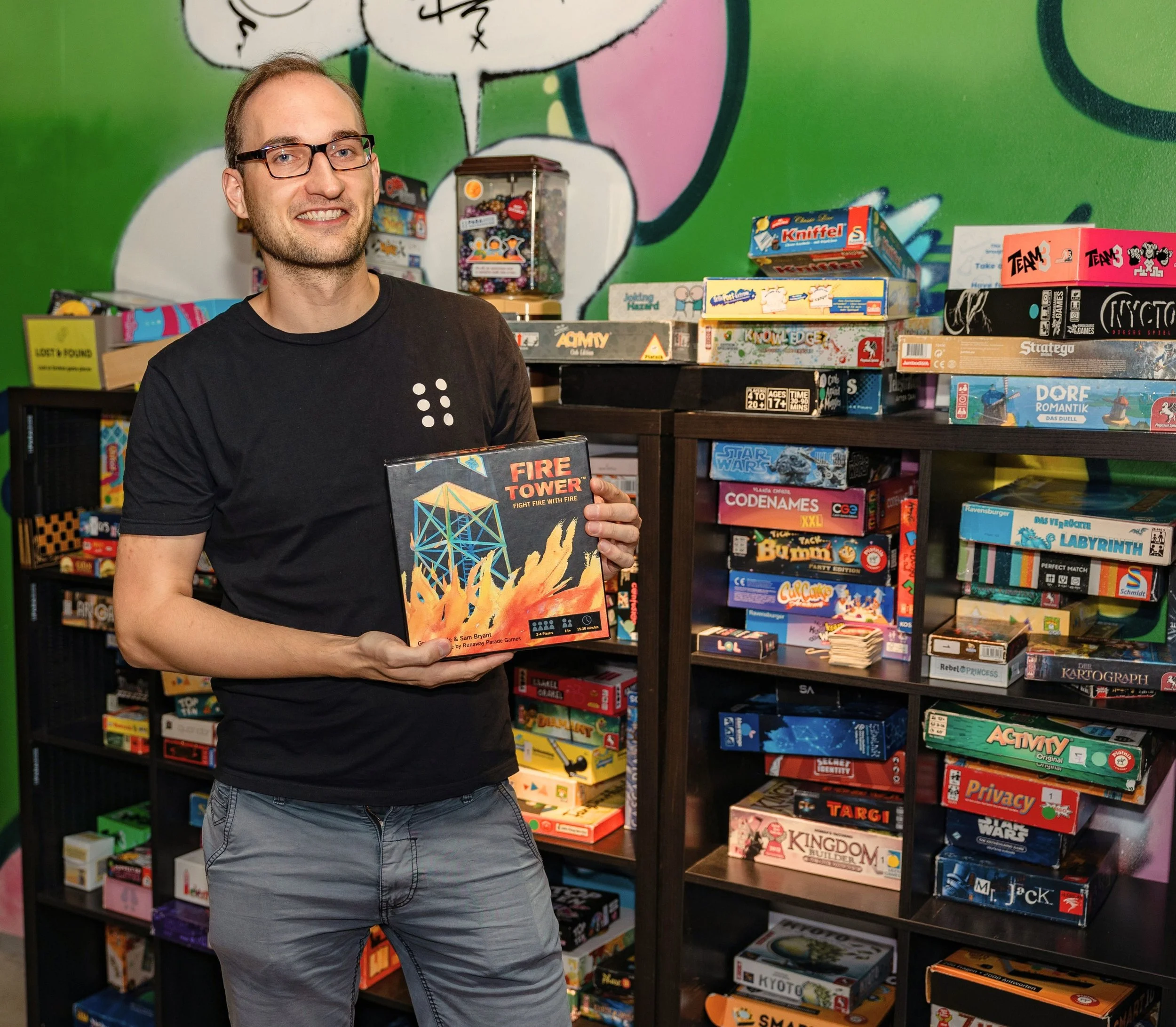 Ein Foto des Finanzvorstands Keziah Loock vor einem Spieleregal. Er hält das Spiel "Fire Tower" in der Hand.