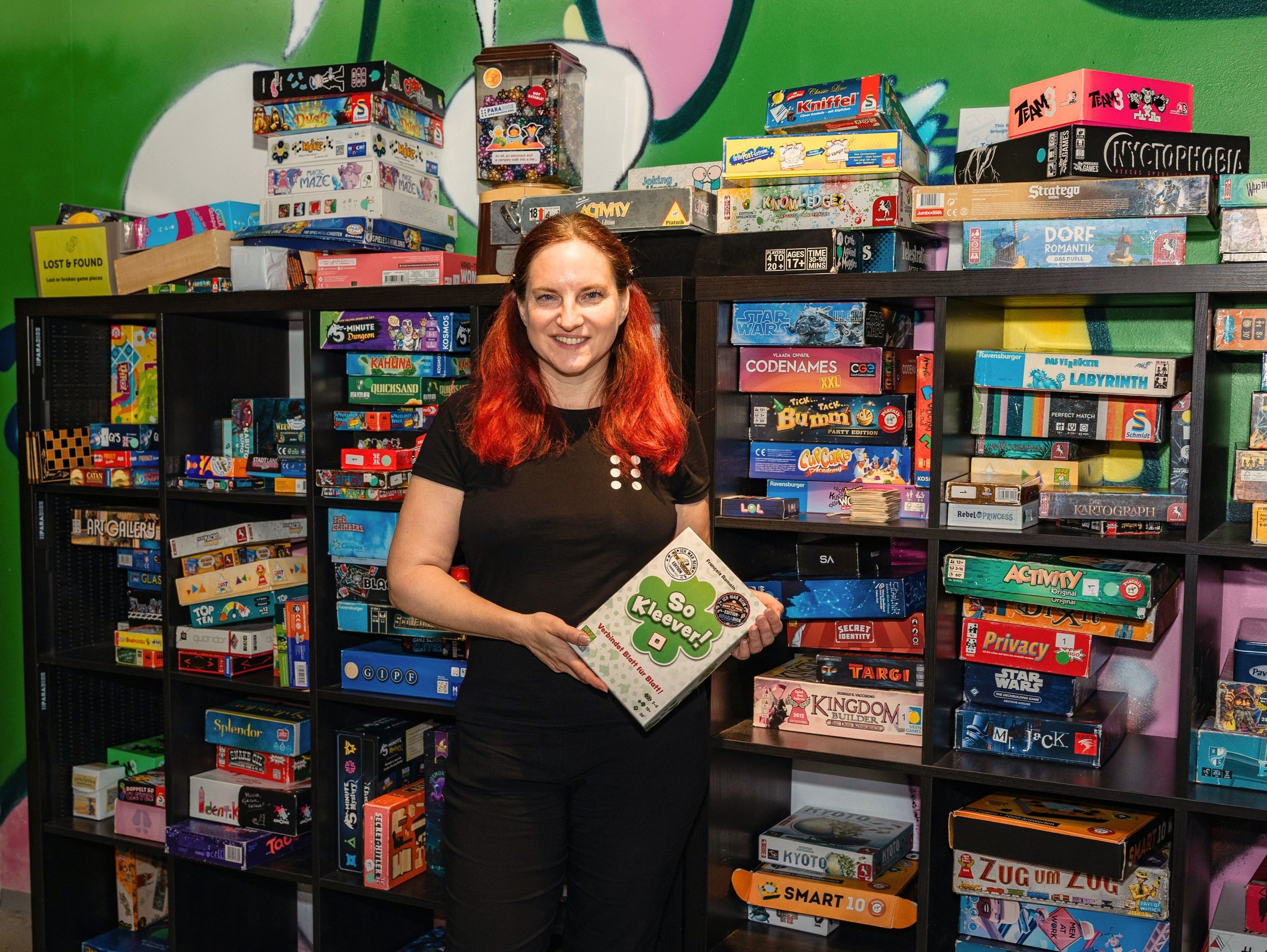 Ein Foto des Vorstandsmitglieds Alice Leinweber vor einem Spieleregal. Sie hält das Spiel "So Kleever!" in der Hand.