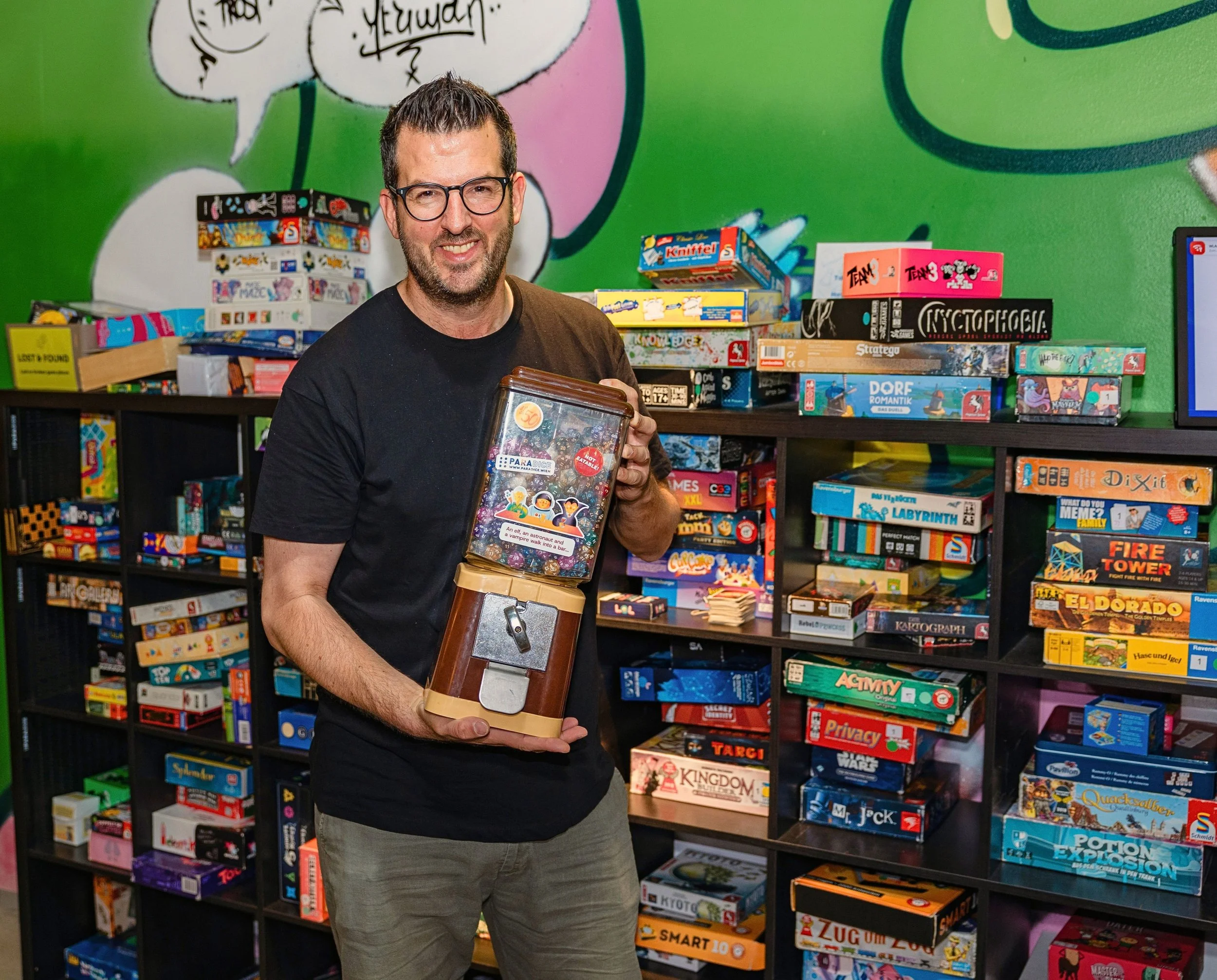Ein Foto des Vorstandsmitglieds Tobias Rosen vor einem Spieleregal. Er hält einen Kaugummispender mit Würfeln in der Hand.