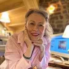 Femme souriante assise dans un café ou un restaurant avec décor en bois, portant une chemise rose clair, posant sa tête sur sa main.