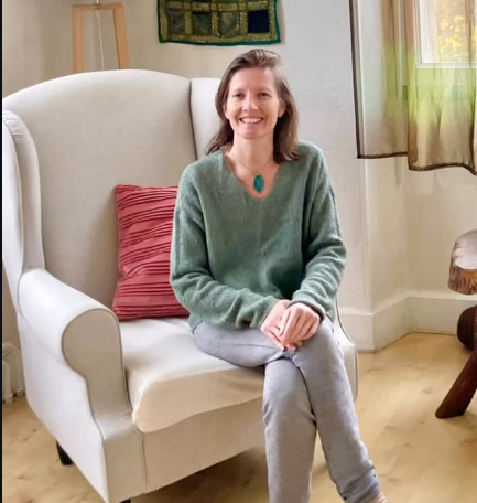 Femme assise sur un fauteuil blanc dans un salon, portant un pull vert et un collier turquoise, souriante.