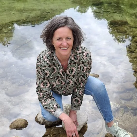 Femme dans une rivière rocheuse, souriante, portant un sweat à motifs et des jeans bleus.