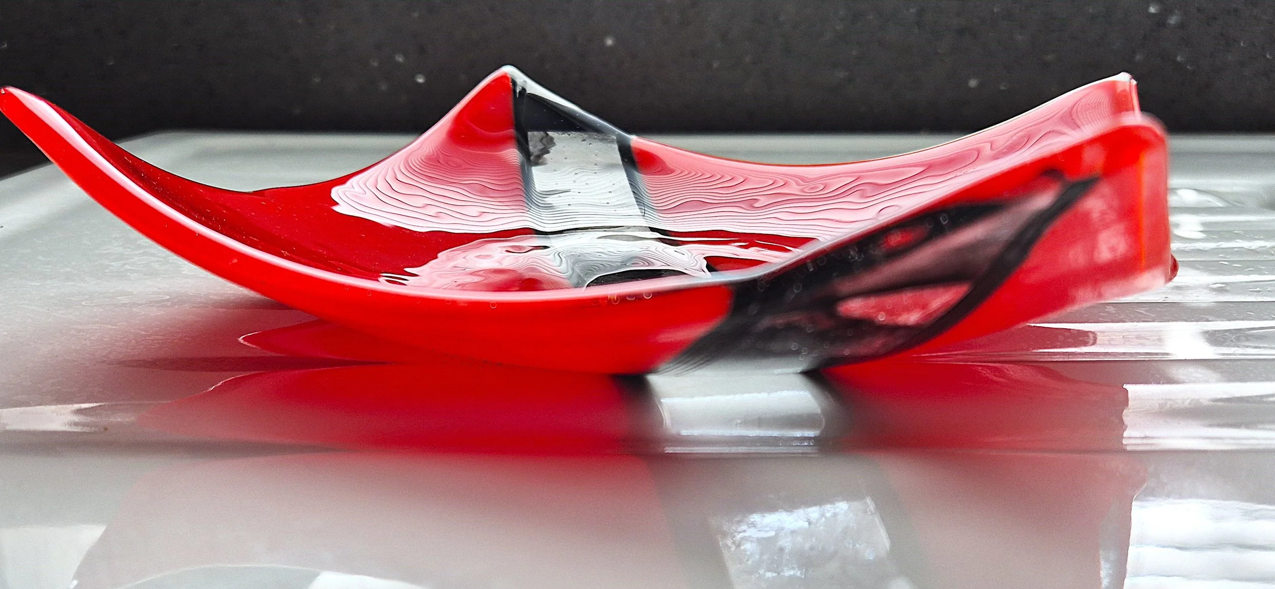 A red and black glass bowl with a wavy, textured surface filled with water, placed on a reflective surface.