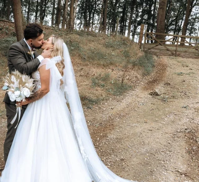 Bride and groom kissing on a rustic path with trees and gate, bride in a wedding dress holding bouquet.