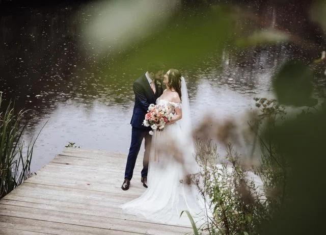 Bride and groom standing on a wooden dock by a lake, surrounded by greenery, embracing and holding a bouquet.