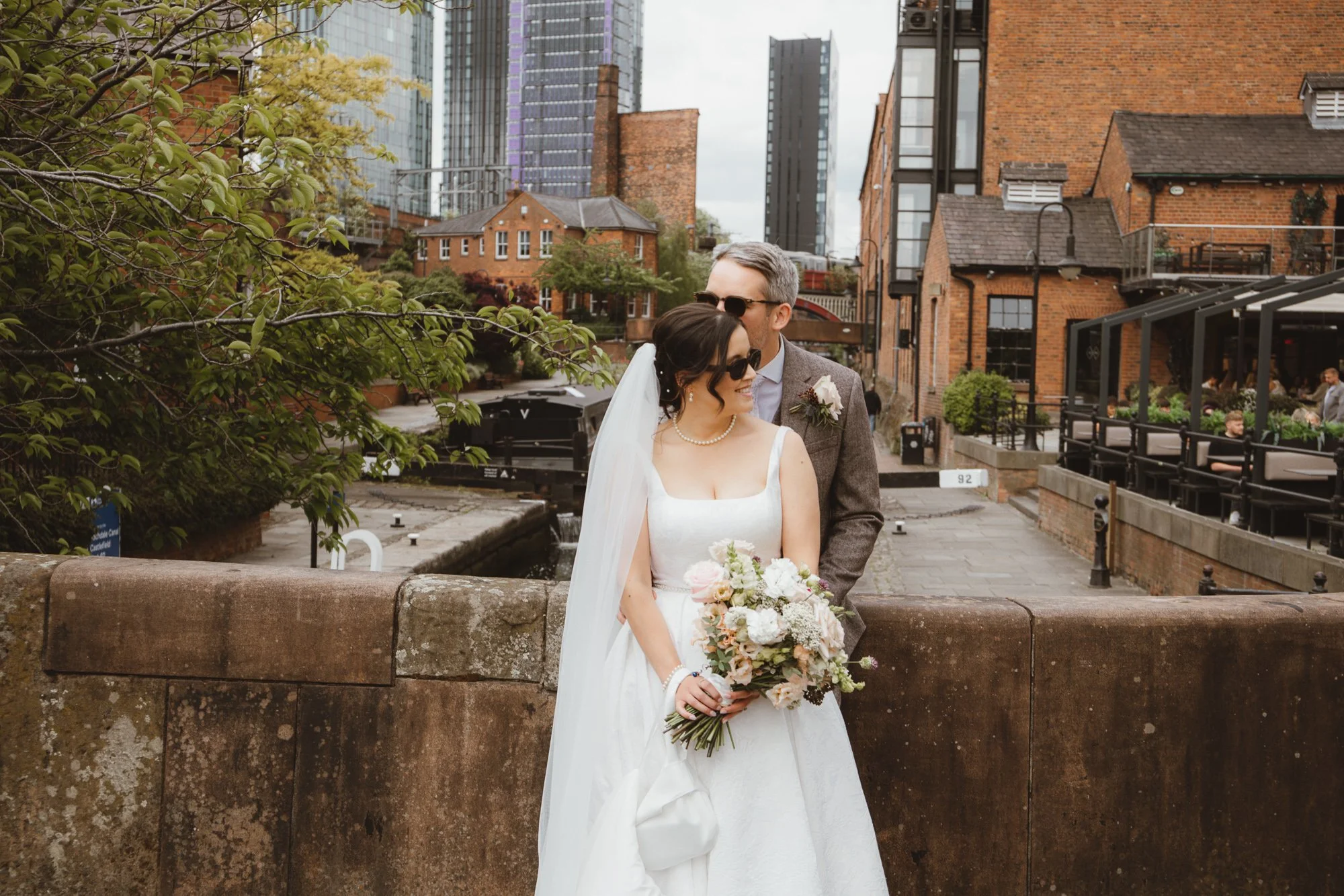 A newlywed couple standing outdoors near a stone wall, with the bride holding a bouquet of flowers and wearing sunglasses, and the groom behind her, also wearing sunglasses, in an urban setting with brick and glass buildings in the background.