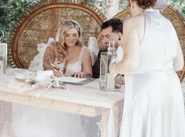 Bride and groom signing a document at a wedding ceremony table with decorative lanterns and flowers, witnessed by a woman in a white dress.