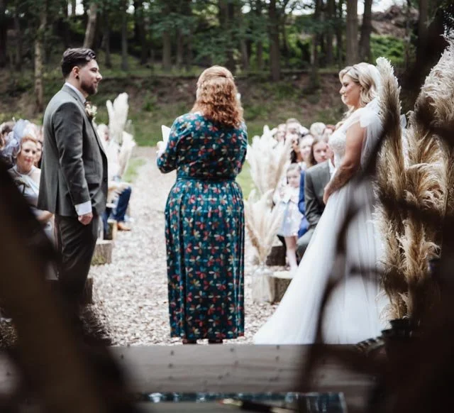 Outdoor wedding ceremony in a wooded area, featuring a bride in a white dress and a groom in a suit, with an officiant standing between them. Guests seated on either side and decorative pampas grass.