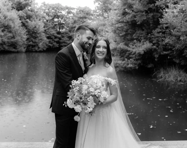 Bride and groom posing by a lake with trees in the background, in black and white.