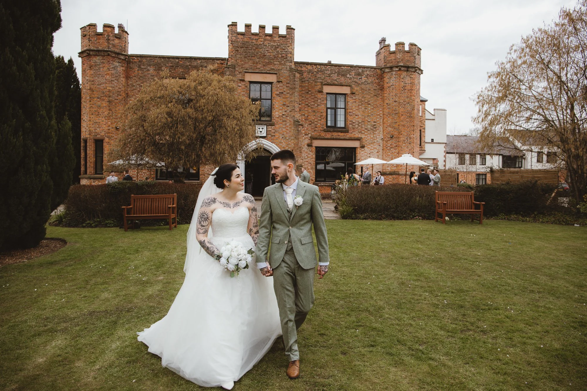 A bride and groom walking on a lawn in front of a brick castle-like building. The bride wears a white wedding dress and holds a bouquet, while the groom wears a gray suit. Guests are in the background near tables with umbrellas.
