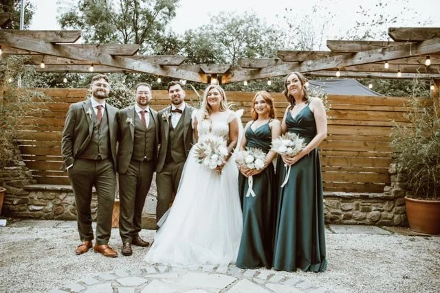 Wedding party posing outdoors under string lights, features Bride in white gown with bouquet, bridesmaids in dark teal dresses with bouquets, and groomsmen in suits with ties, standing in front of wooden fence and greenery backdrop.