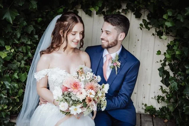 Bride and groom sitting together, bride holding a bouquet, surrounded by greenery.
