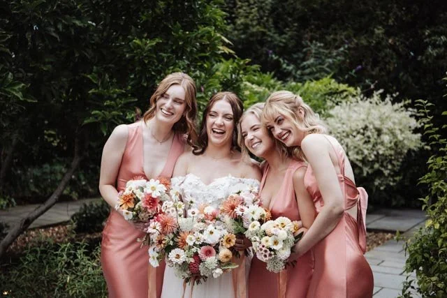 Bride with three bridesmaids in pink dresses holding bouquets, smiling in a garden setting.