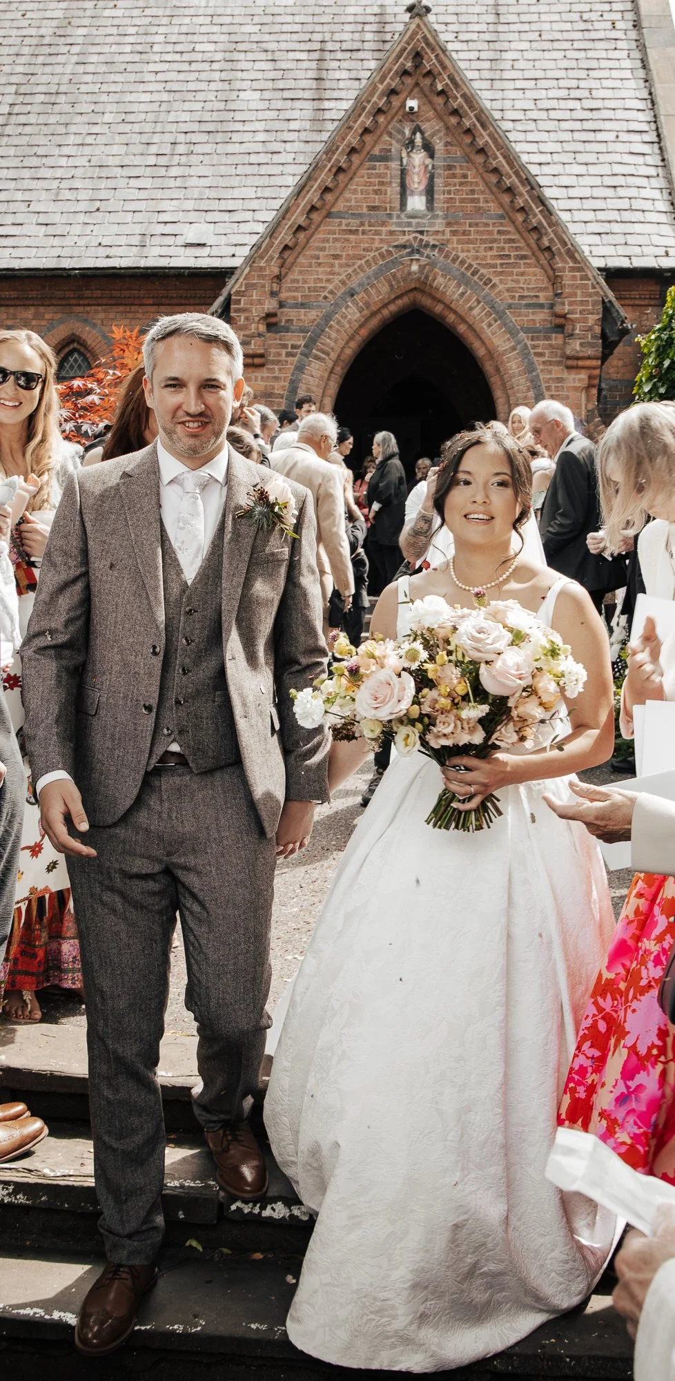 A bride and groom standing outside a brick church, holding hands, with guests in the background. The bride is wearing a white dress and holding a bouquet of flowers. The groom is dressed in a gray suit with a boutonniere. The church exterior is made of red bricks with a small shrine or religious image above the entrance.