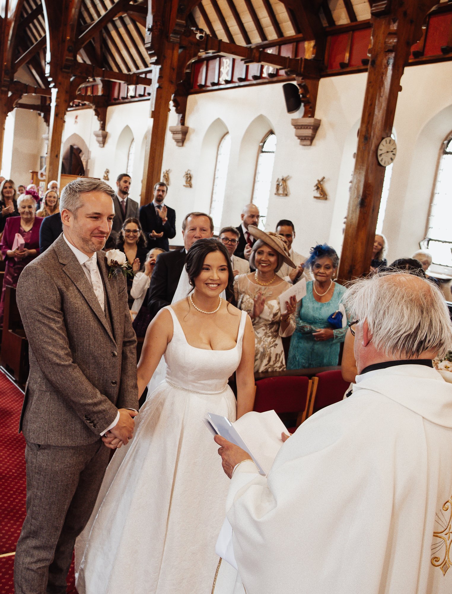 A wedding ceremony taking place inside a church with wooden beams and arched windows. The bride and groom stand before the officiant, holding hands, while guests are seated and standing, clapping and smiling.