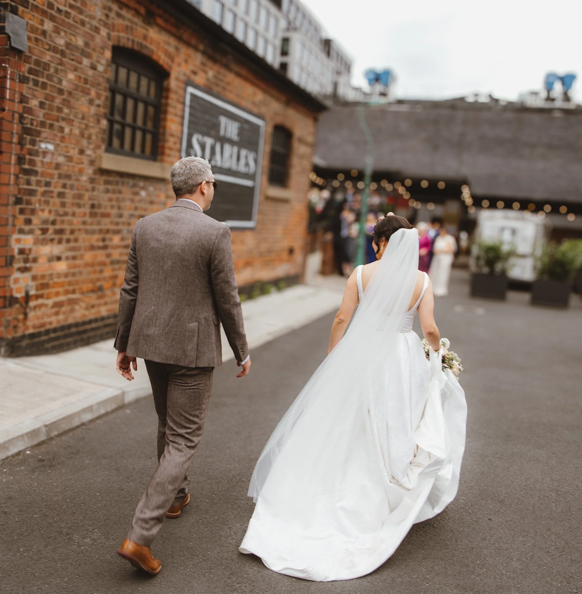 Bride and groom walking outdoors near a brick building, bride in a white wedding dress with a long train and veil, groom in a brown suit, in a city setting with others in the background.
