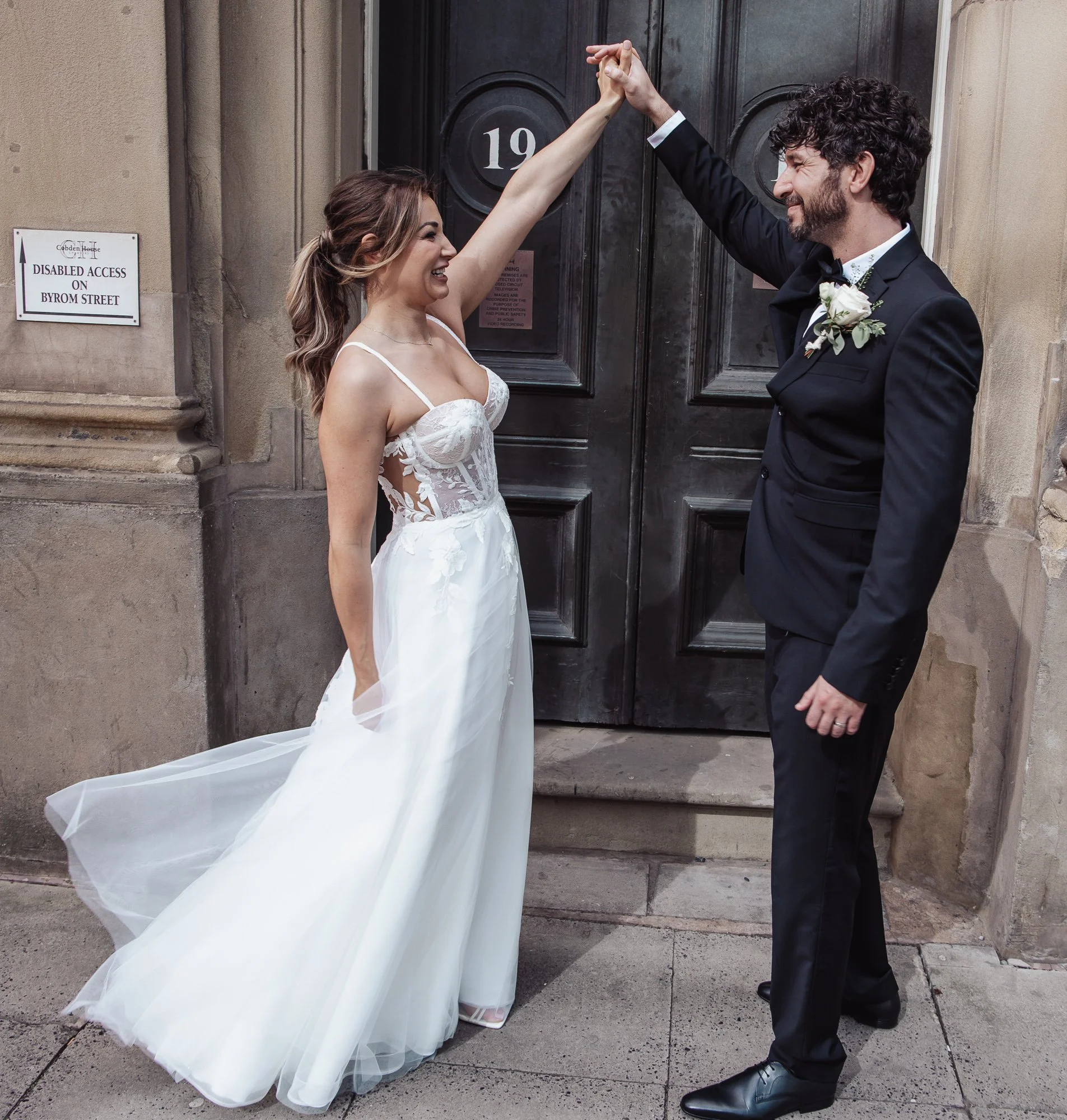 A bride and groom dancing in front of a door marked with the number 19. The bride is wearing a white wedding dress, and the groom is in a black suit with a boutonniere. There is a sign indicating disabled access nearby.