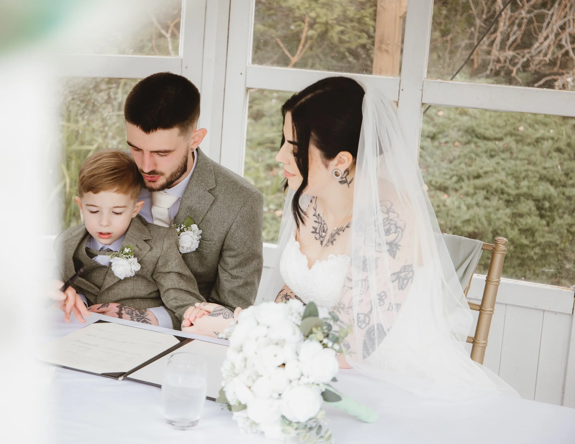 A wedding scene with a groom, a bride, and a young boy, all wearing formal attire. The groom and boy are in gray suits with white flower boutonnieres. The bride is in a white dress with tattoos visible, wearing a veil, sitting at a table with a document and bouquet.