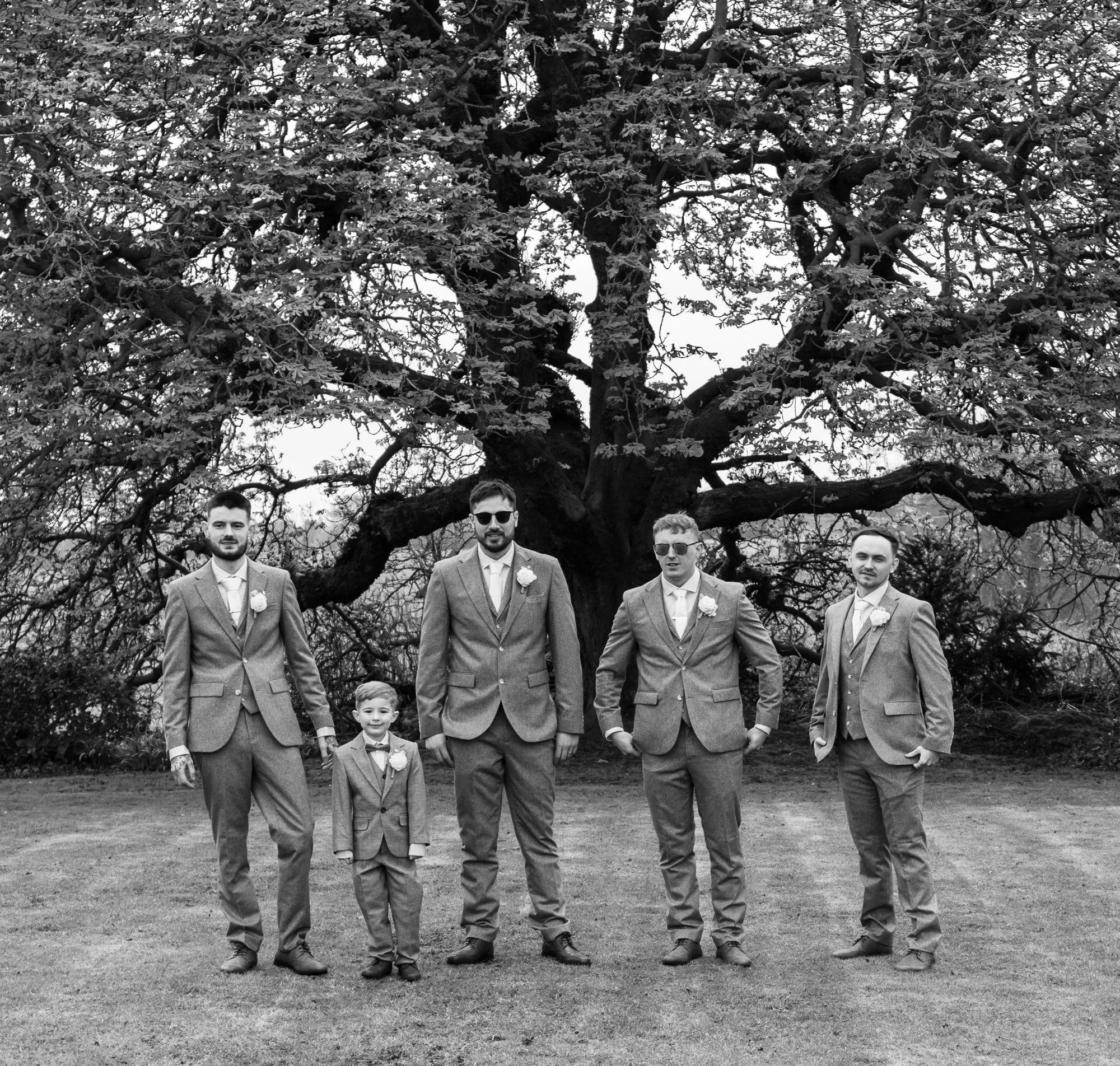 Five men, including one child, dressed in suits and standing in front of a large tree. The setting is outdoors on a grassy area. The image is black and white.