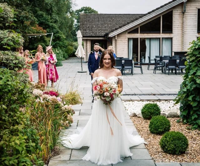 Bride walking down a garden path holding a bouquet, followed by bridesmaids and a groomsman near a modern house with a patio.