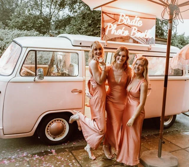 Three women in matching pink dresses stand smiling in front of a vintage pink van with a "Birdie Photo Booth" sign.