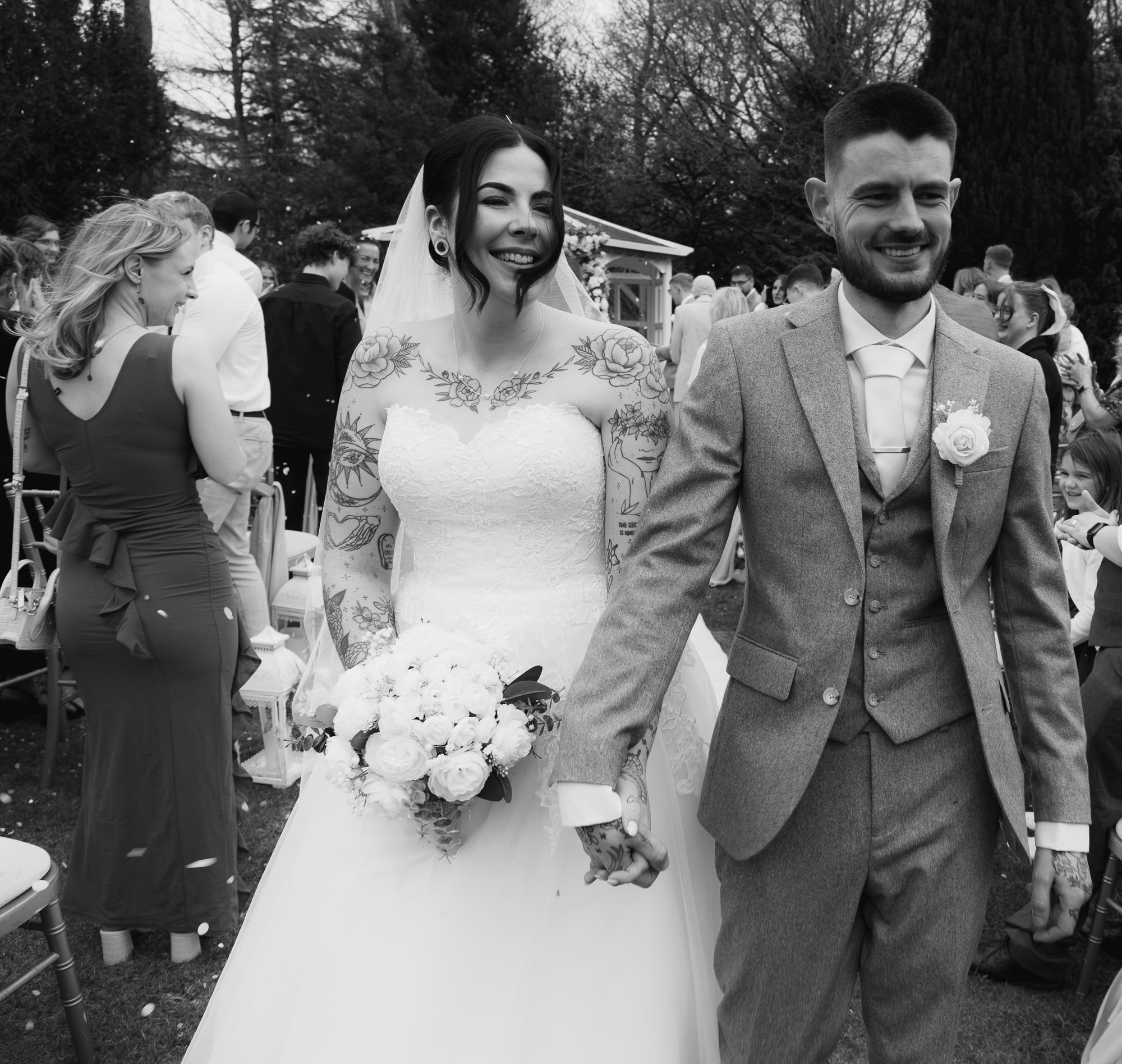 A newlywed couple walking together outdoors, the bride in a lace wedding dress holding a bouquet of white flowers, and the groom wearing a gray suit with a boutonnière. They are surrounded by guests at an outdoor venue, smiling and celebrating.
