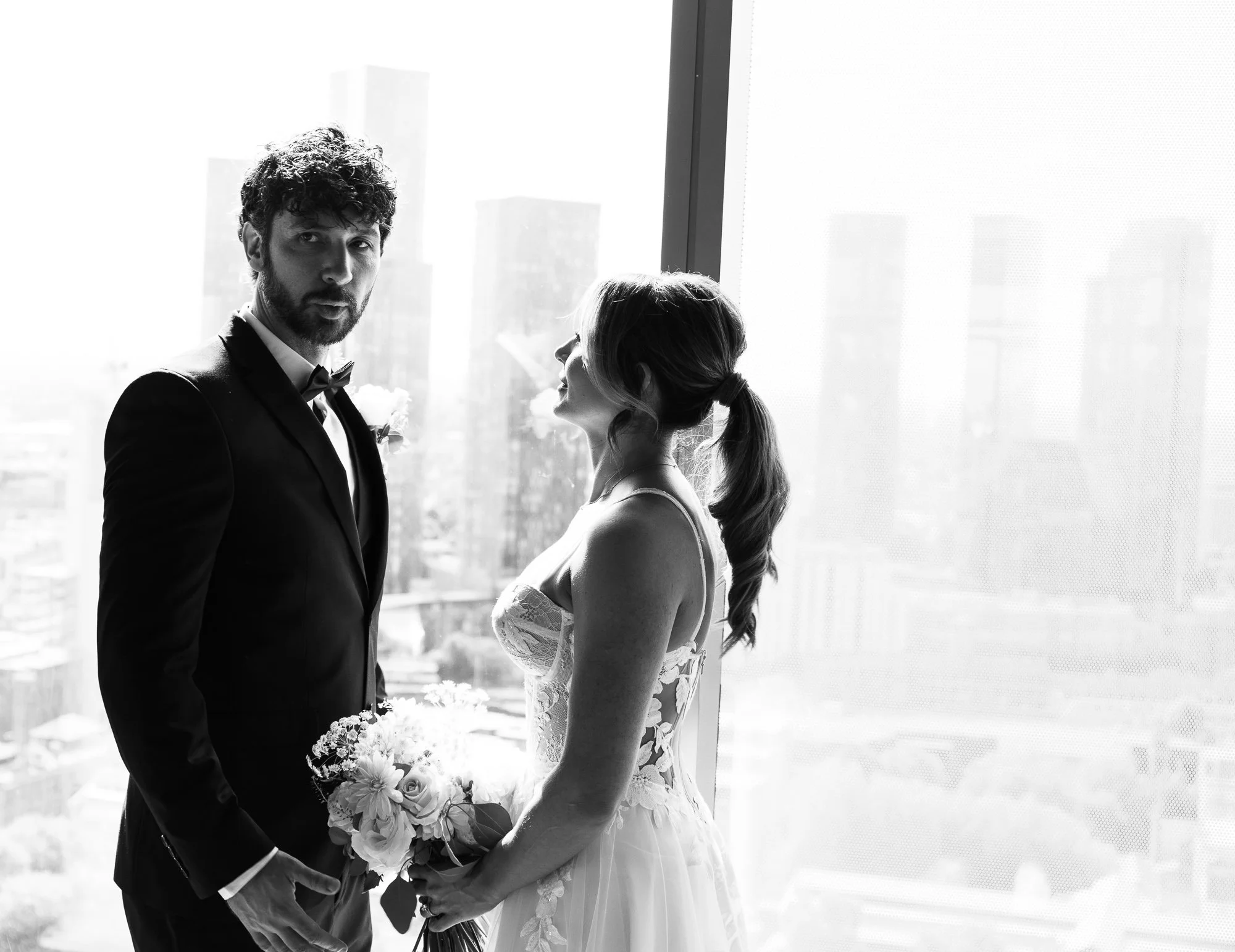 Black and white photo of a bride and groom standing by a large window with a cityscape in the background. The groom is wearing a suit and bow tie, and the bride is wearing a wedding dress and holding a bouquet.