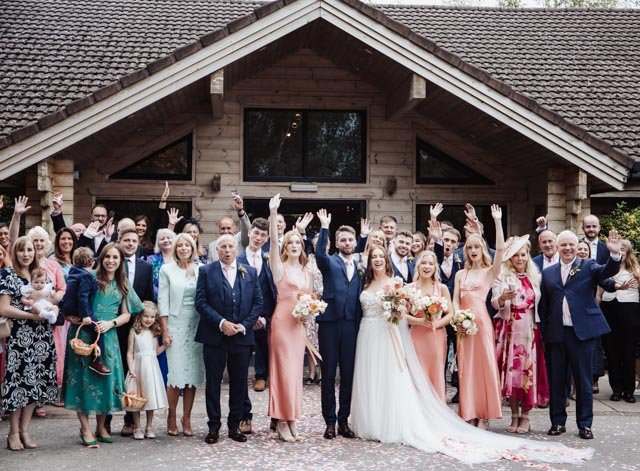 Group wedding photo featuring a newlywed couple and guests in front of a wooden building. The bride is in a white dress, and the groom is in a blue suit. Bridesmaids in pink dresses, and several guests are waving hands in celebration.