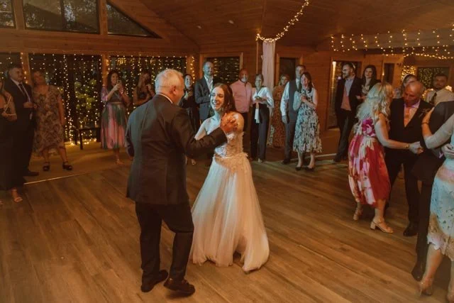A bride and an older man dancing at a wedding reception, surrounded by guests. The venue is warmly lit with string lights.