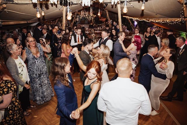 People dancing at a lively indoor wedding reception with dim lighting and string lights.