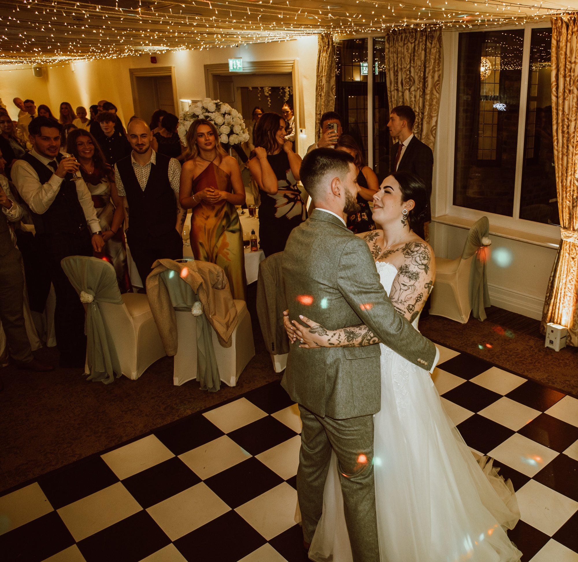 Couple dancing at a wedding reception with guests watching on a checkered floor and festive lights in the background.