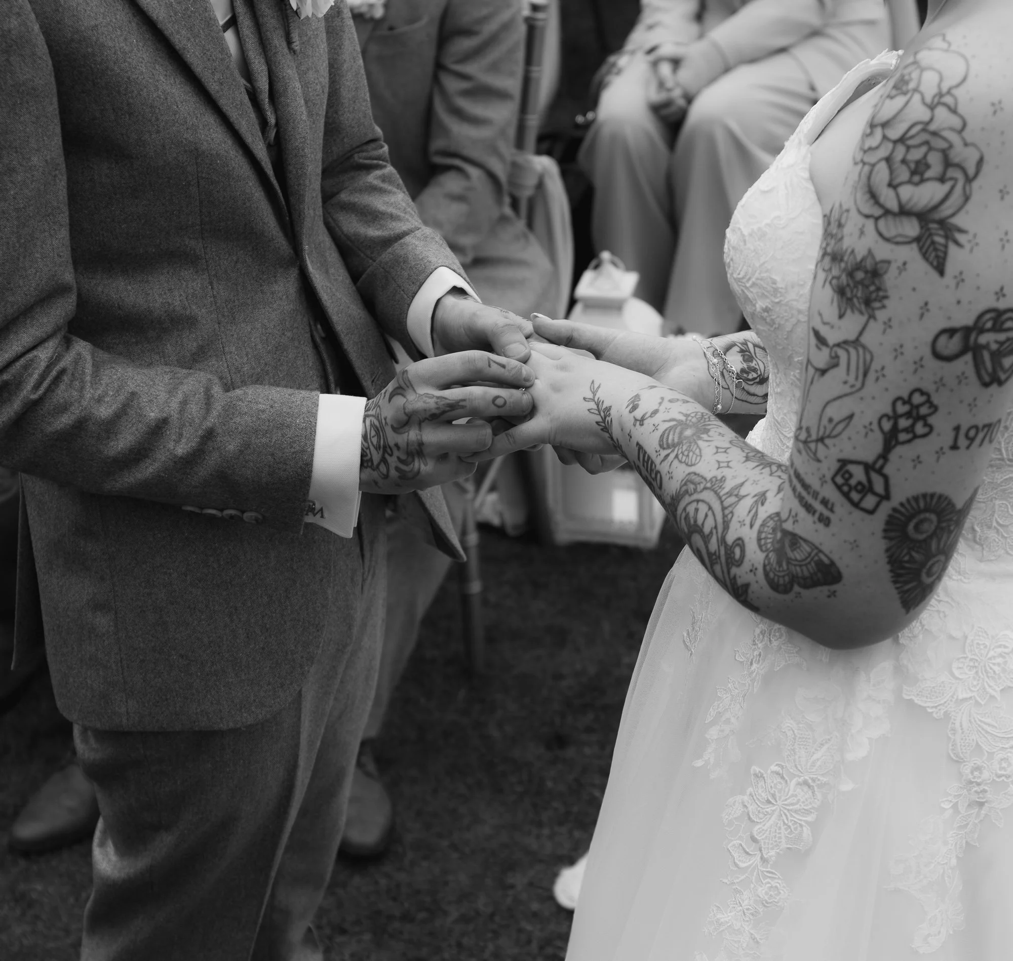 Black and white photo of a wedding ceremony where a person in a suit places a ring on a tattooed person's finger.