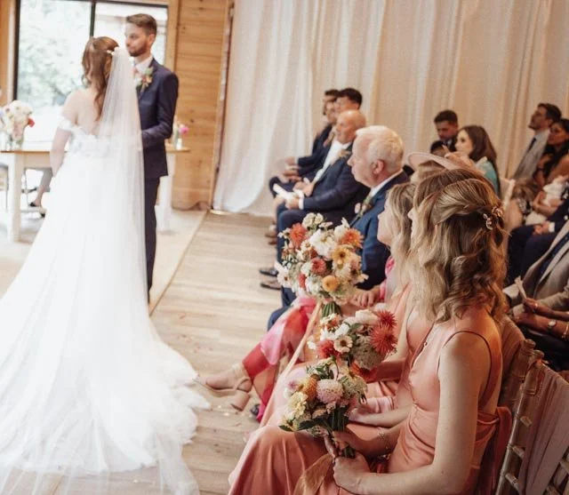 Wedding ceremony with bride, groom, bridesmaids, and guests seated in a wooden venue.