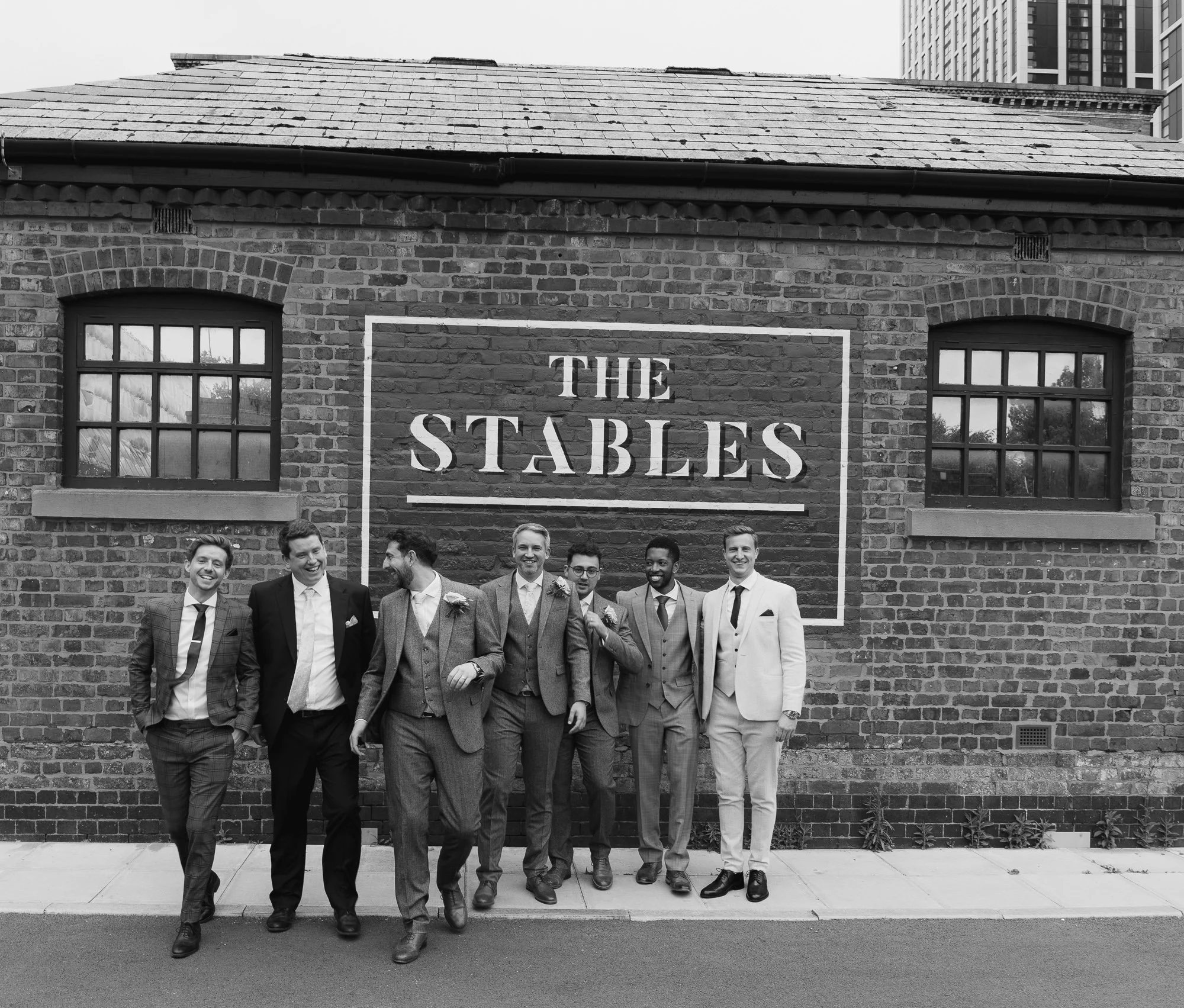 Group of seven men dressed in suits walking and smiling in front of a brick wall with a sign that reads 'The Stables'