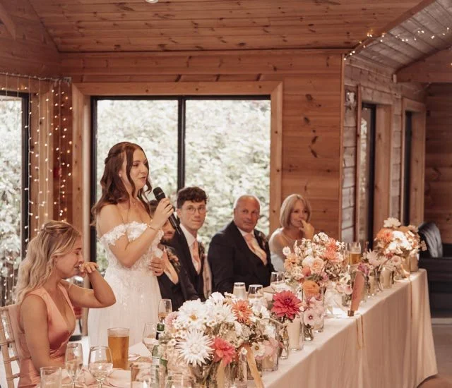Bride giving speech at wedding reception with guests seated at a table, decorated with flowers, inside a wooden venue.