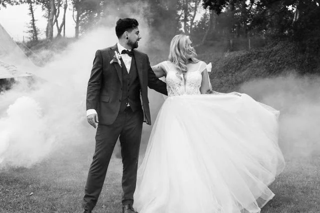 Bride and groom in formal attire standing on grass with smoke in the background, black and white photo.