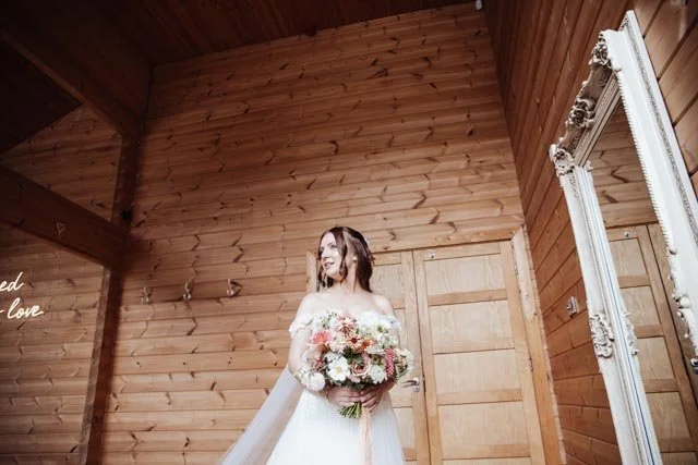 Bride in a wooden room holding a bouquet of flowers near a large ornate mirror.