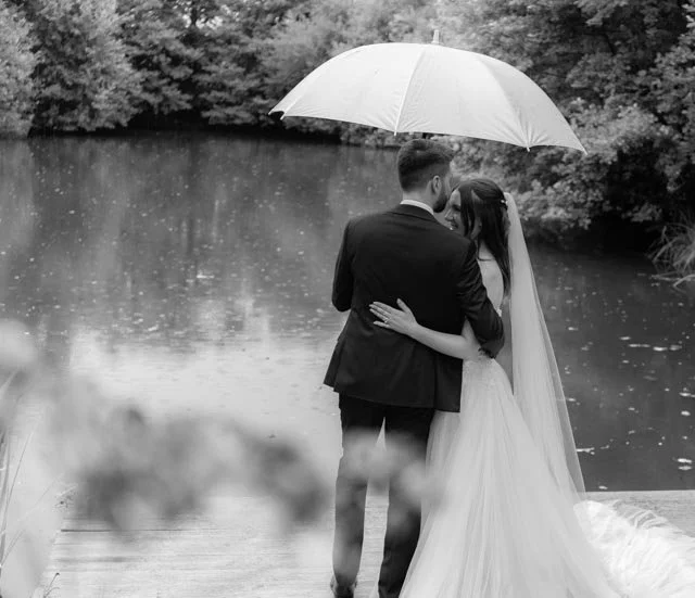 Bride and groom holding an umbrella by a lake, in black and white.