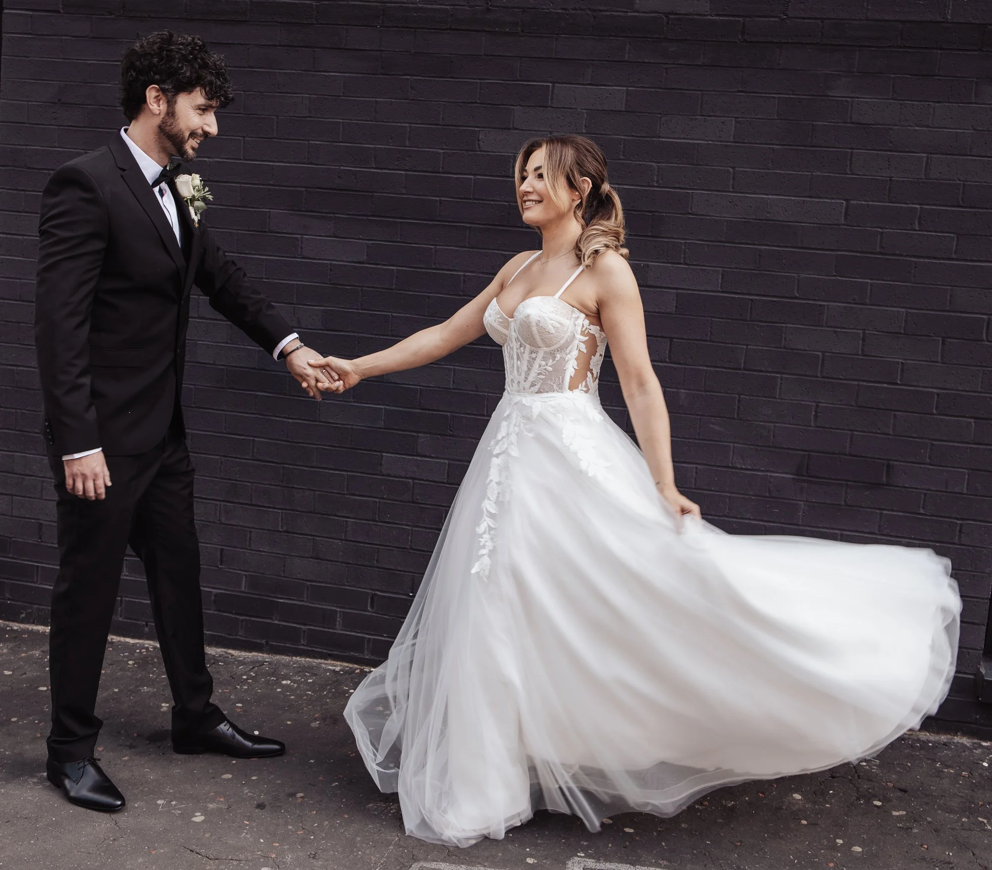 Bride in lace wedding dress holds hands with groom in black suit against dark brick wall.