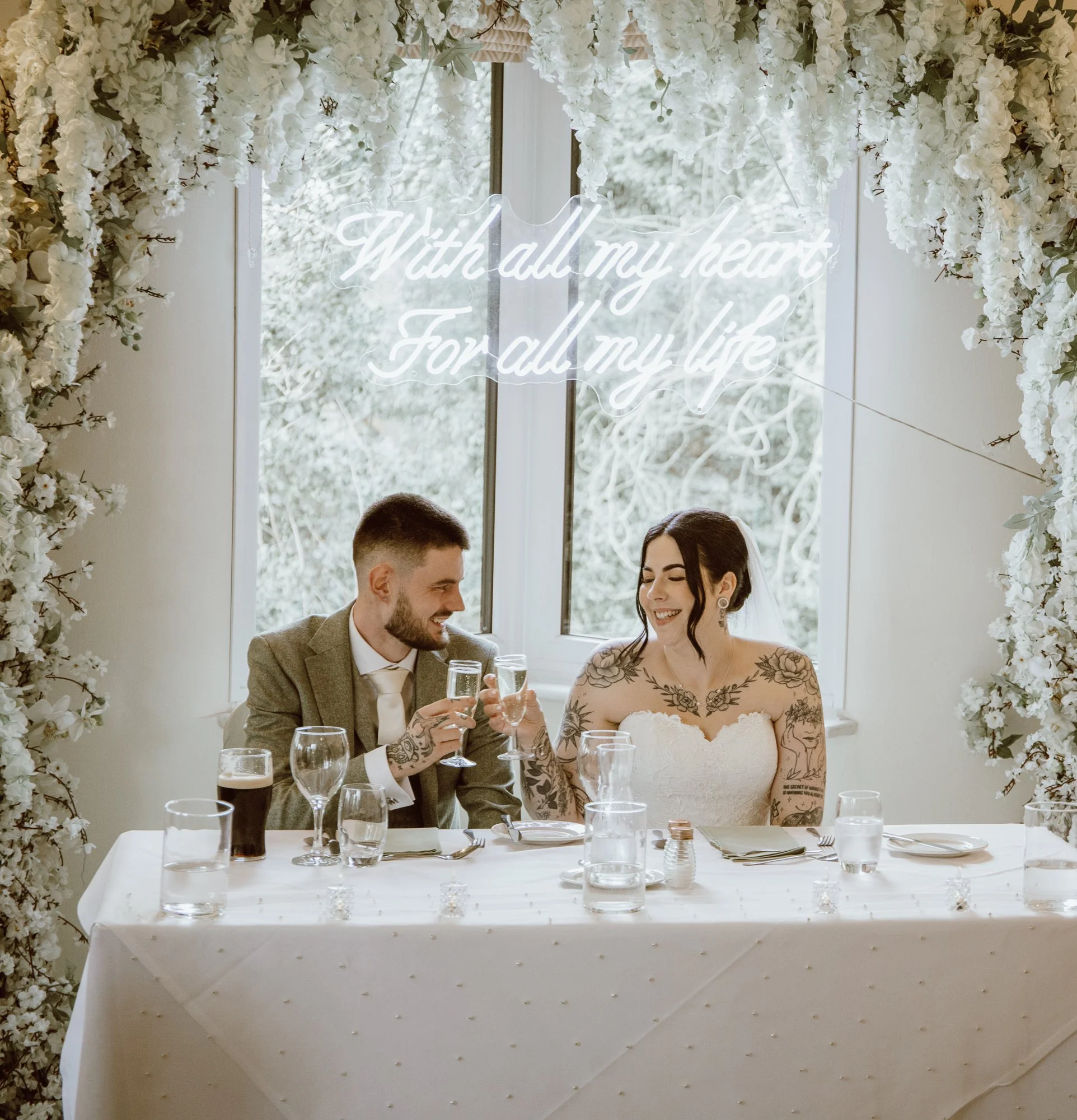 Bride and groom clinking glasses at a wedding table, surrounded by floral decor, with a neon sign reading 'With all my heart, For all my life' in the background.