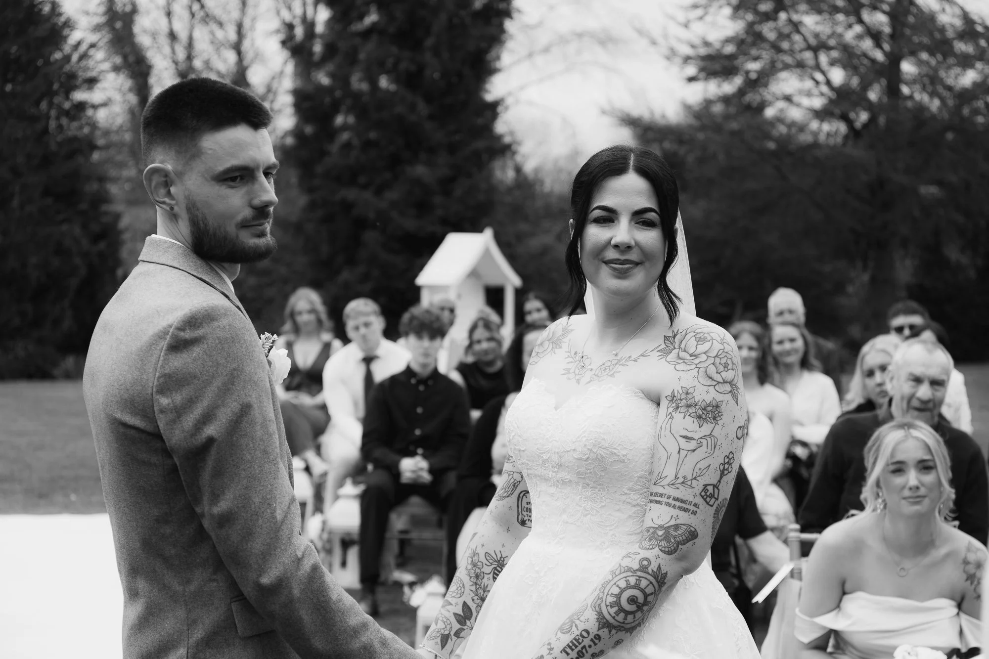 Bride and groom at outdoor wedding ceremony with guests seated behind them.