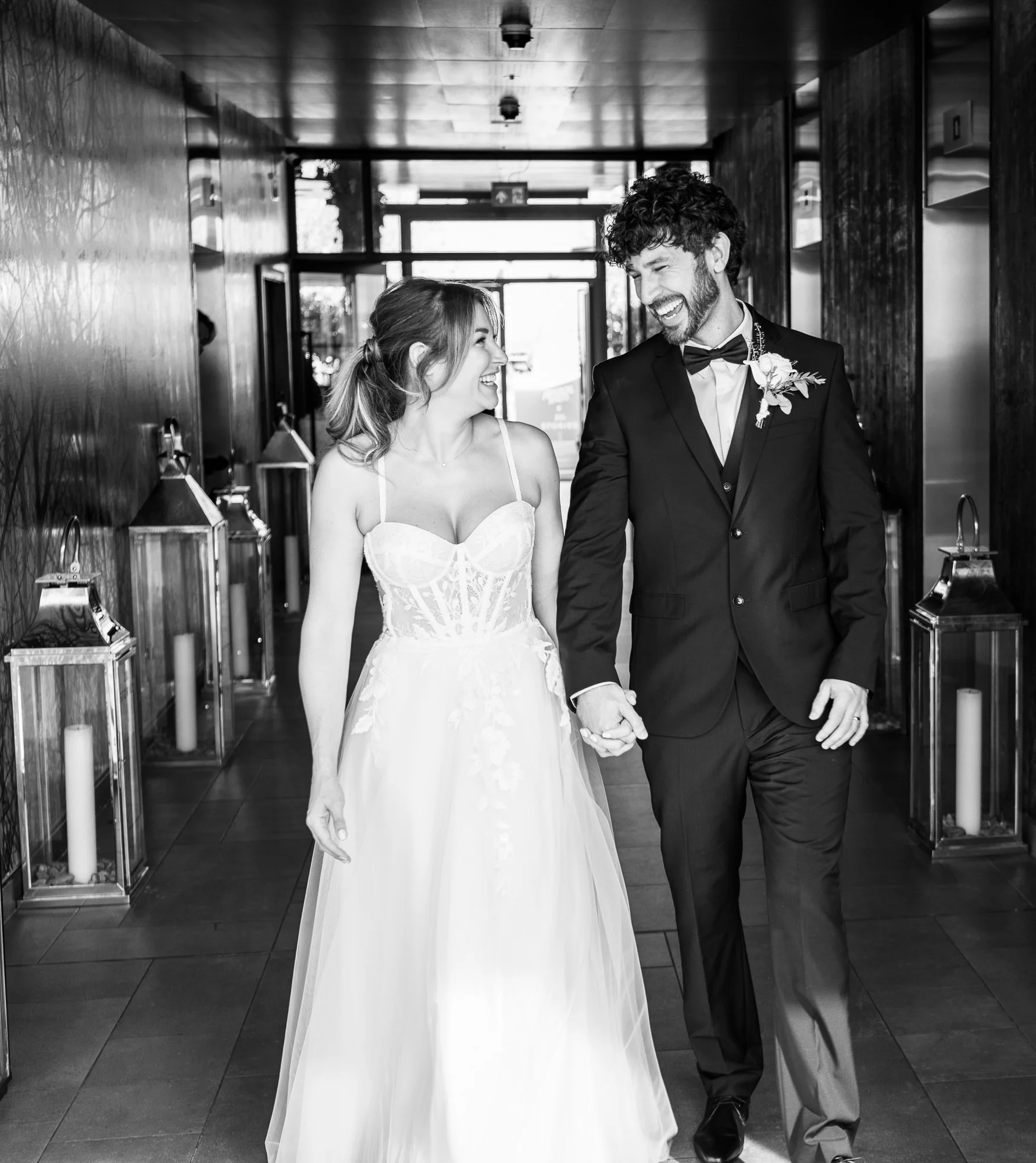 Bride and groom walking hand in hand, smiling at each other, in a hallway with decorative lanterns, black and white photo.