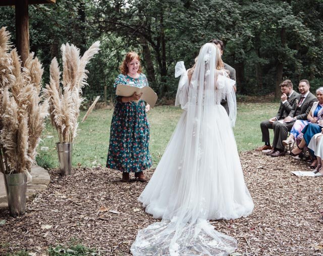 Outdoor wedding ceremony with bride, groom, officiant, and seated guests. Bride in white dress and veil facing groom. Officiant holding papers, standing among pampas grass decorations. Guests seated on the right.