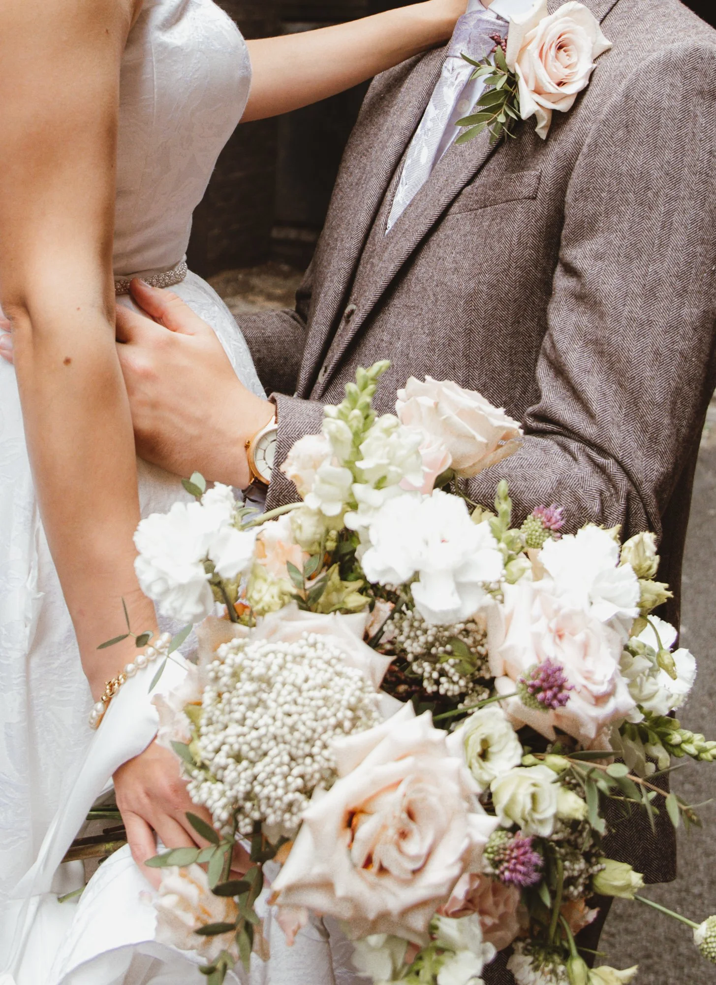 Close-up of a bride and groom at their wedding, with the bride holding a bouquet of white and blush flowers and the groom wearing a gray suit with a pink rose boutonniere, embracing each other.