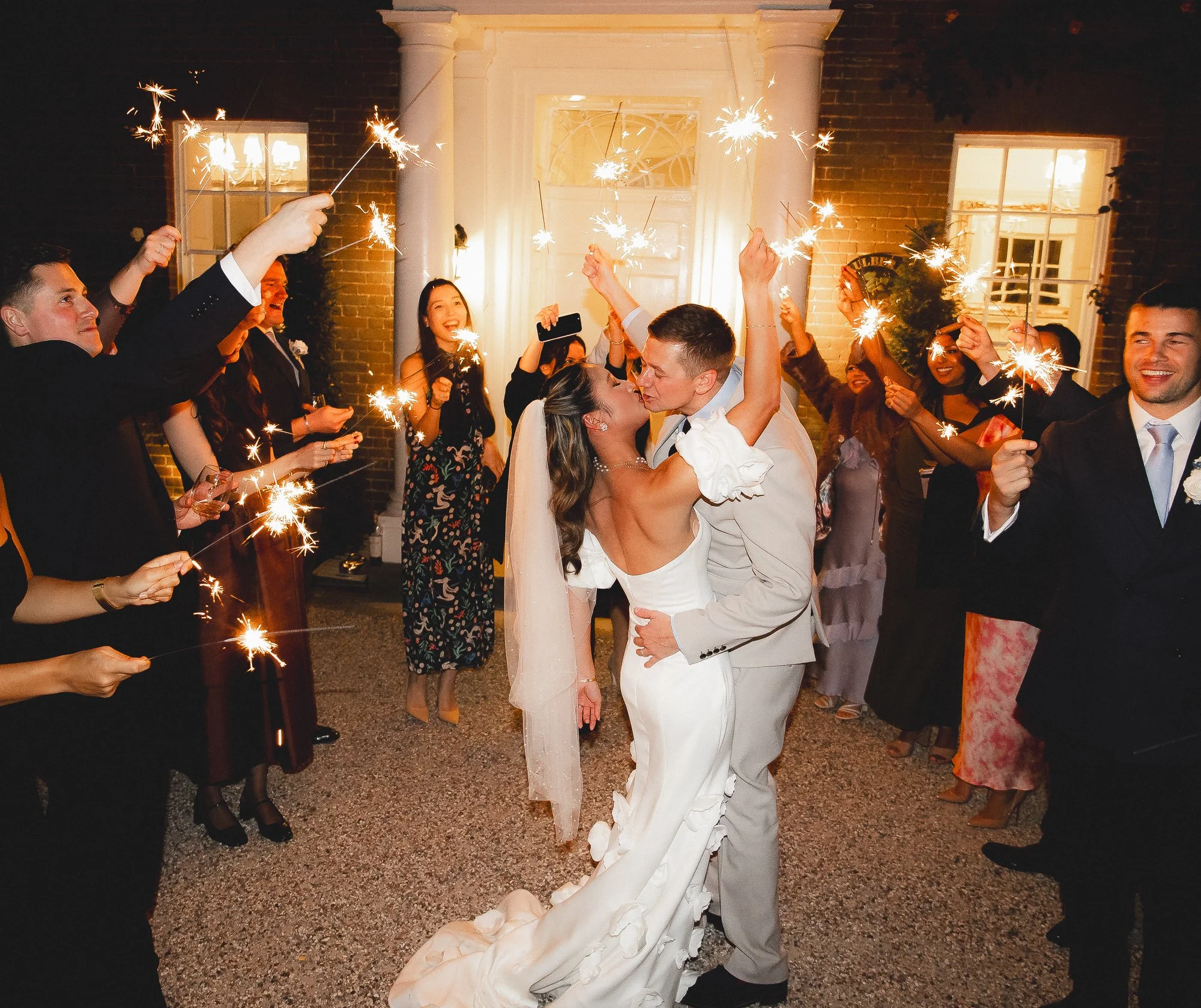 A newlywed couple in wedding attire sharing a kiss and dancing surrounded by friends holding sparklers at night outside a building with white columns.