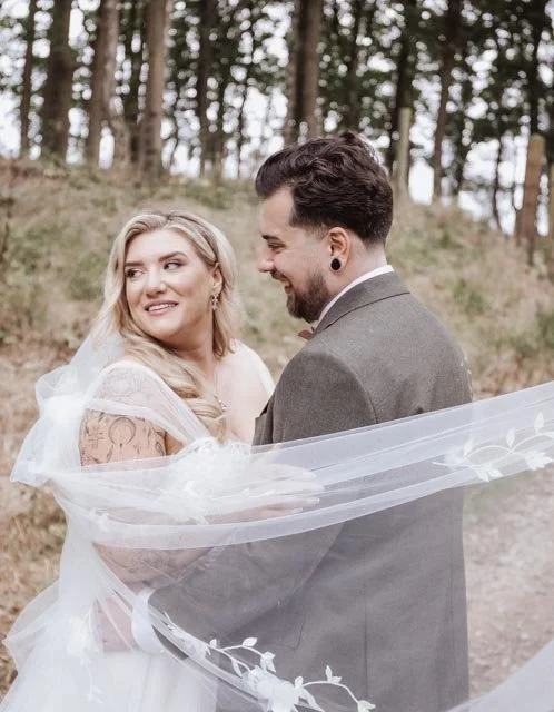 Bride and groom smiling outdoors, bride's veil flowing, forest background.