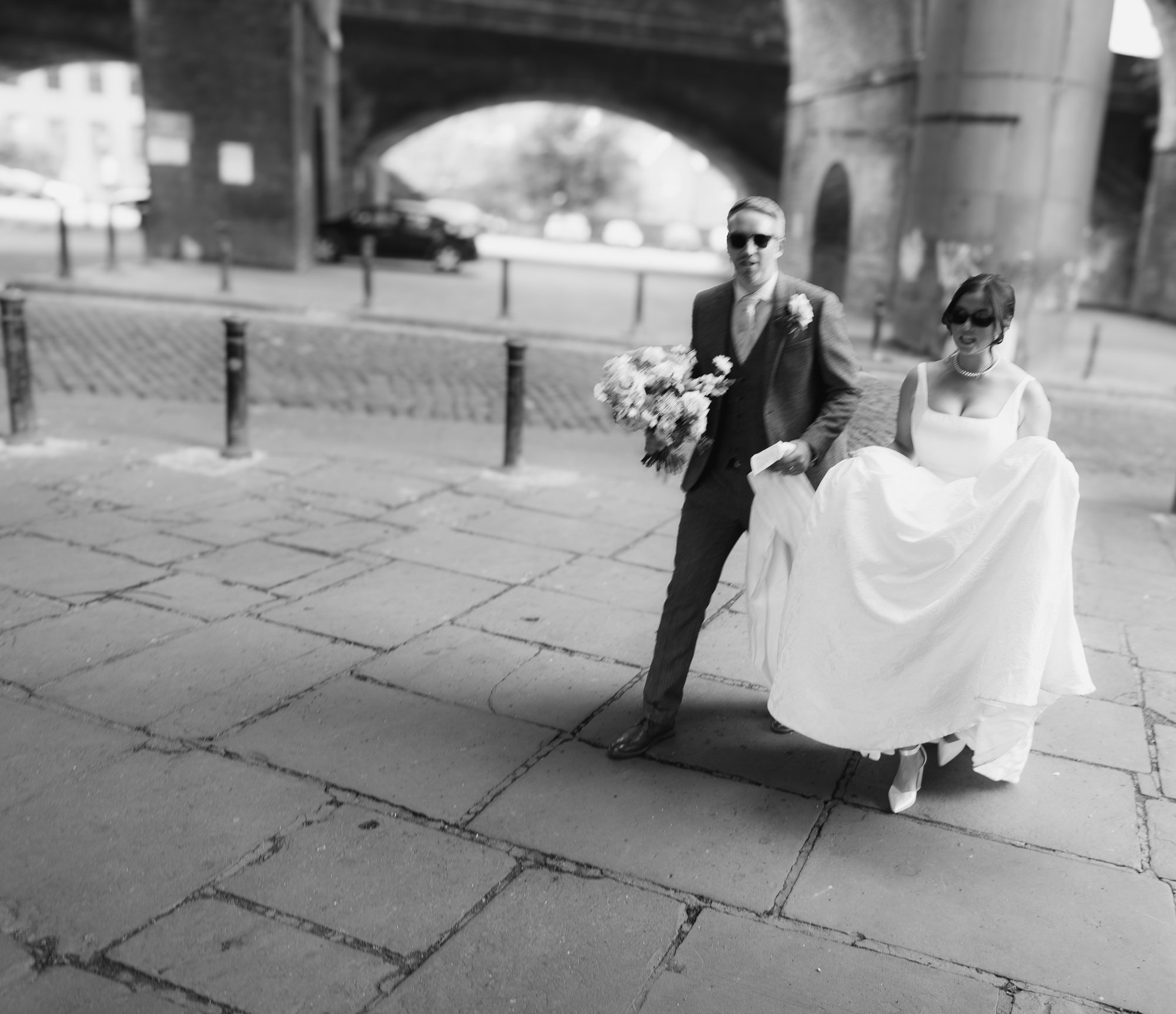 A black and white photo of a groom carrying a bouquet of flowers and a bride wearing a wedding dress walking together outdoors under a bridge.