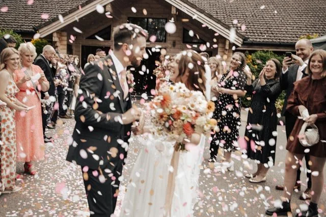 Bride and groom surrounded by guests throwing confetti outside a wooden building, with bride holding a bouquet of white and orange flowers.